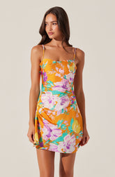 A woman is wearing a sleeveless dress with thin spaghetti straps. The dress features a bright orange background with a large floral pattern in shades of purple, pink, yellow, and teal. The dress has a fitted bodice and a slightly gathered detail on one side, creating a subtle drape effect.